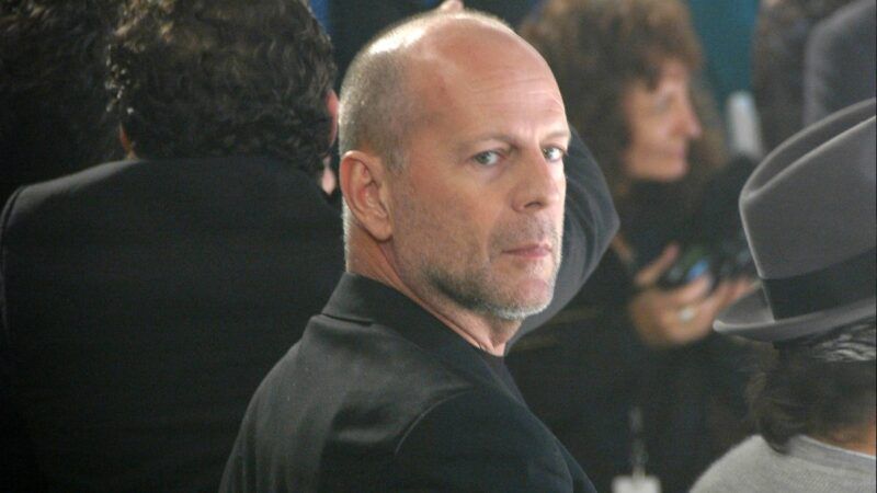 Bruce Willis viste un traje negro y mira a la cámara por encima del hombro a través de la multitud.