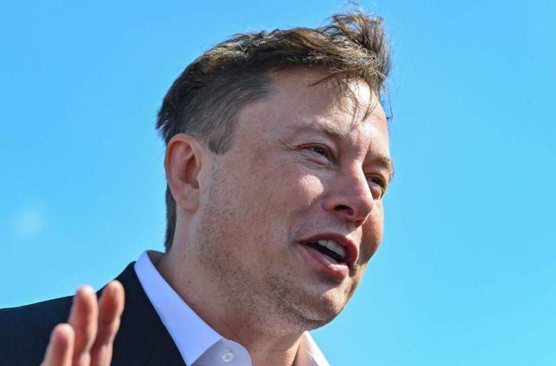 Elon Musk purtând un costum și ridicând mâna.