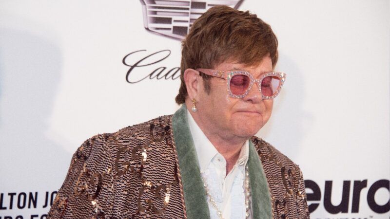 Elton John na rdeči preprogi nosi bleščeč suknjič na belem ozadju