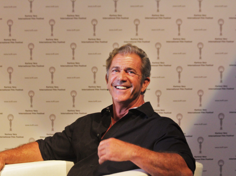 Mel Gibson sentado en una silla, sonriendo, vestido con una camiseta negra