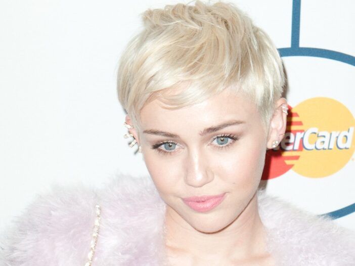 Miley Cyrus má na červenom koberci oblečený páperový ružový kabát cez krátke biele šaty