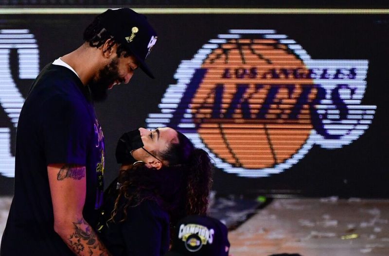 Anthony Davis con un sombrero negro y una camiseta abrazando a su novia, que lleva una máscara negra. Están parados en una cancha de baloncesto frente al logo de los Lakers.