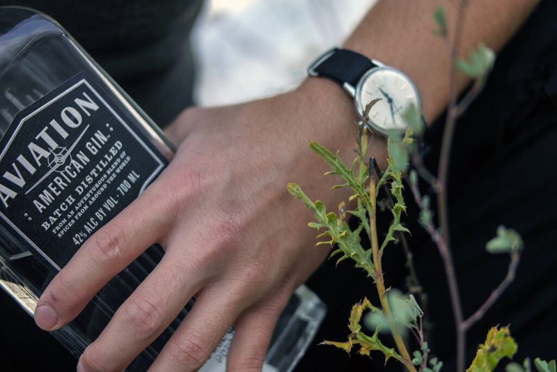 Una mano con un reloj de pulsera, sosteniendo una botella de Aviation American Gin.