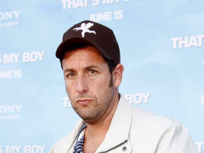 Adam Sandler fa il broncio e indossa una giacca bianca con un cappellino da baseball nero.