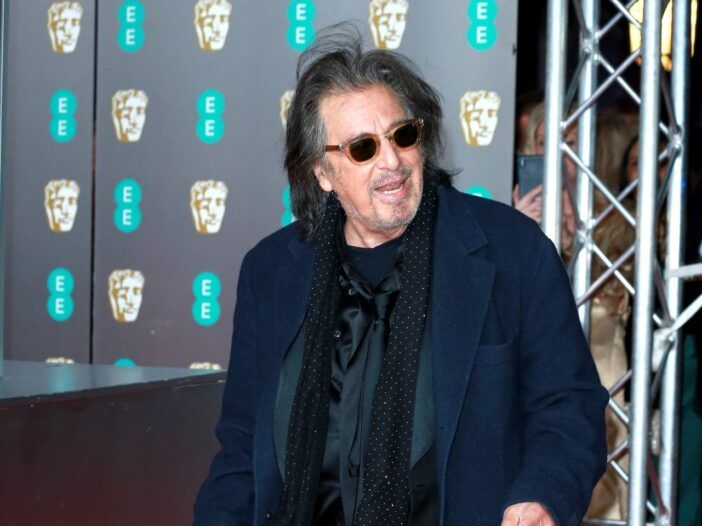 Al Pacino de pie con la boca abierta y vestido con una chaqueta azul, gafas de sol negras y una bufanda negra.