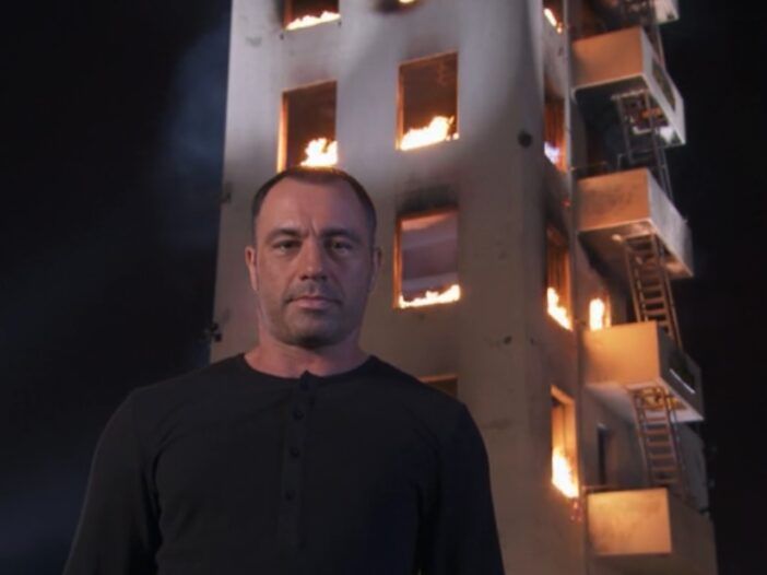 Joe Rogan seisab põleva hoone ees, seljas must särk ja tühi näoilme.