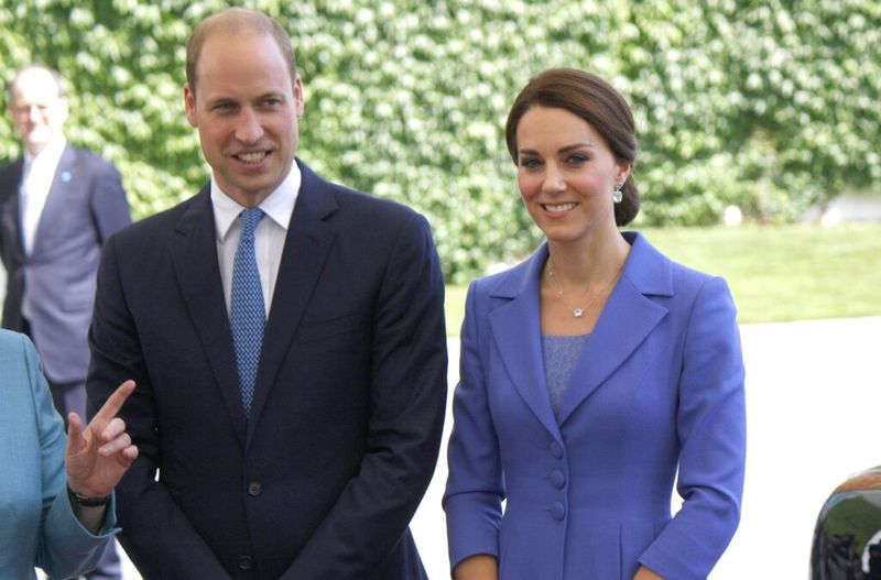 Princas Williamas tamsiu kostiumu su mėlynu kaklaraiščiu stovi kartu su Kate Middleton mėlynu kostiumu