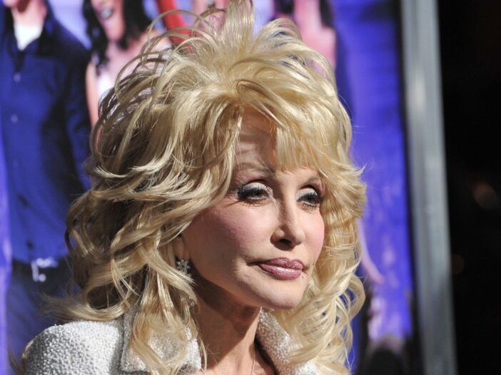 Dolly Parton on pukeutunut valkoiseen haalaripukuun violetilla taustalla