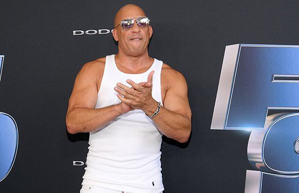 Vin Diesel con una camiseta sin mangas blanca, vaqueros blancos y gafas de sol, aplaudiendo.