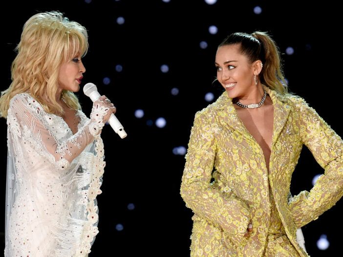 Dolly Parton vľavo v bielom, vystupujúca na pódiu s Miley Cyrus, vpravo v zlatom