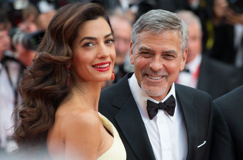 Amal Clooney sonríe por encima del hombro mientras se para con George Clooney, quien
