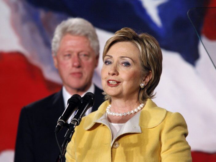 Hillary Clintonová v popredí oblečená v žltom nohavičkovom kostýme, Bill Clinton v pozadí sa pozerá, ako Hillary hovorí na pódiu.