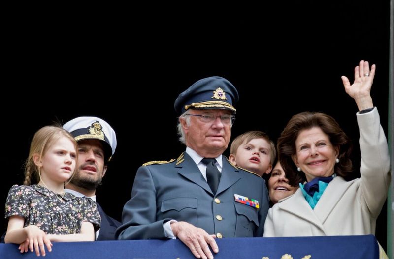 El Rey de Suecia vestido con un traje militar, mirando hacia la multitud junto con su esposa e hijos saludando.
