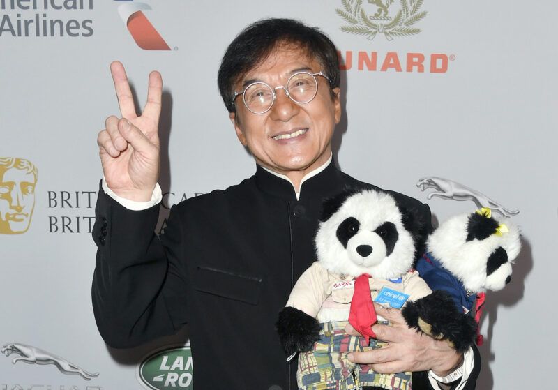 Jackie Chan naeratab mustas riietuses ja hoiab käes pandamänguasju