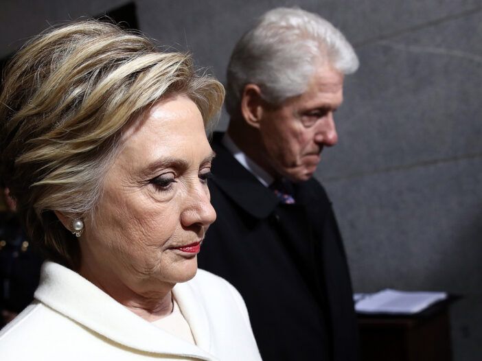 Hillary Clinton en primer plano luciendo adusta, Bill Cljnton ligeramente desenfocado en el fondo.