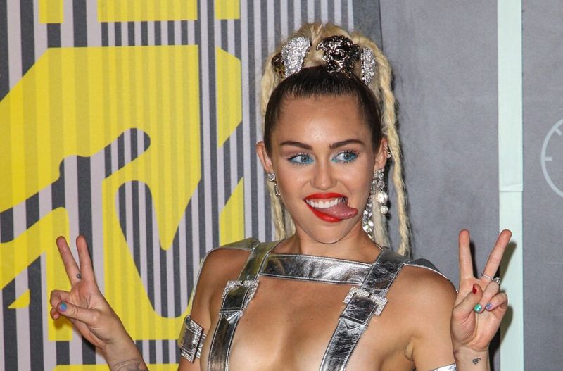 Miley Cyrus sosteniendo dos signos de paz y sacando la lengua.