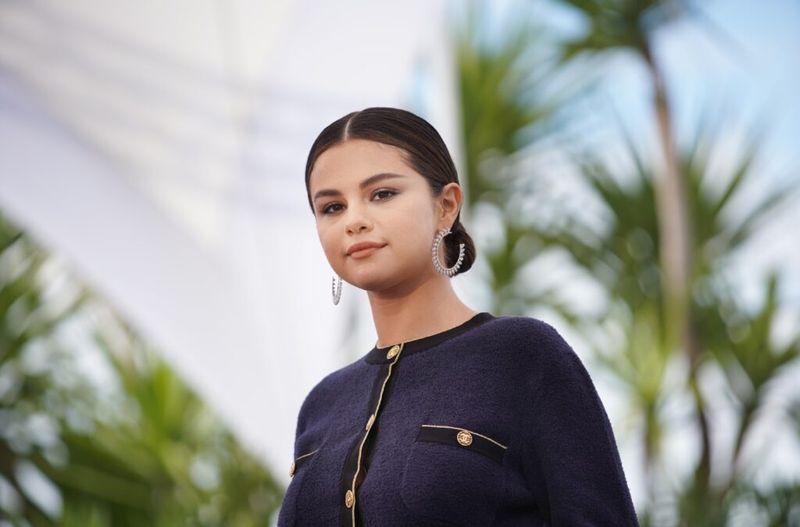 Selena Gomez sonriendo y vistiendo una chaqueta azul marino y dorada mientras está de pie frente a las palmeras.