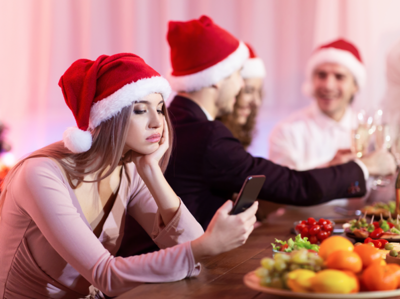 Fiesta de Navidad aburrida. Chica aburrida con smartphone celebrando la Navidad