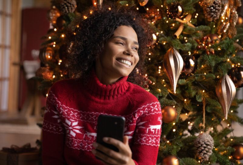 Usmievavá žena vo vianočnom svetri, ktorý drží smartfón pred vianočným stromčekom.