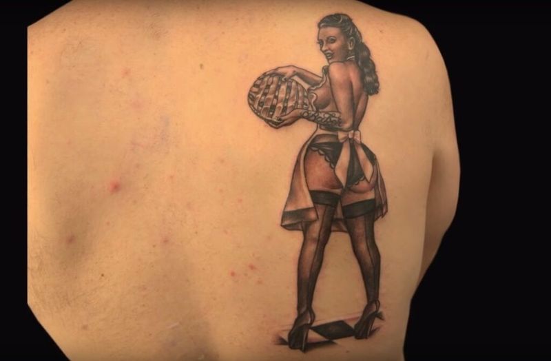 melns un pelēks muguras tetovējums ar sievieti, kas paceļ pīrāgu zeķēs un priekšautu, autors Tatu Baby