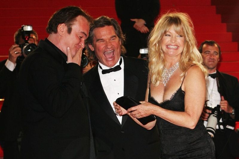 Kventins Taratino melnā uzvalkā, Kurts Rasels smokingā un Goldija Hona melnā kleitā smaida un smejas