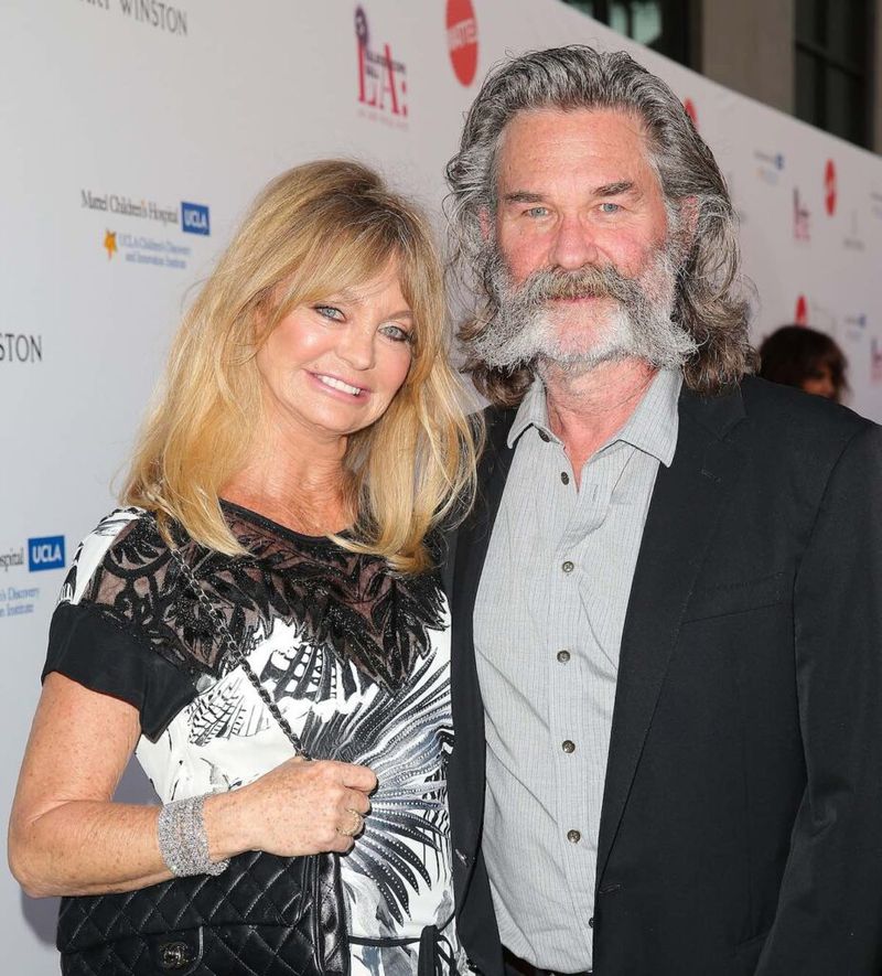 Kurt Russell med spisst skjegg og dress smiler med Goldie Hawn i svart og hvit kjole