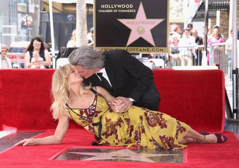Kurt Russell i dress kneler og kysser en tilbakelent Goldie Hawn i en gul kjole foran stjernen hennes