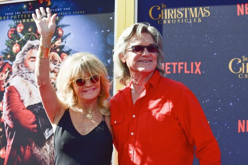 Kurt Russell i rød skjorte smiler ved siden av en vinkende Goldie Hawn kledd i svart