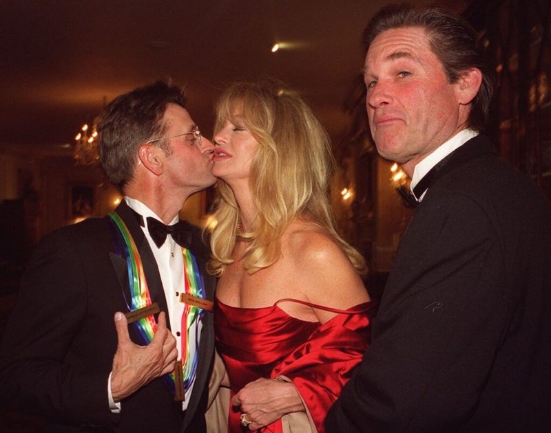 Mikhail Baryshnikov v smokingu poljubi Goldie Hawn (rdeča obleka) na lice, medtem ko Kurt Russell v smokingu dviguje obrvi