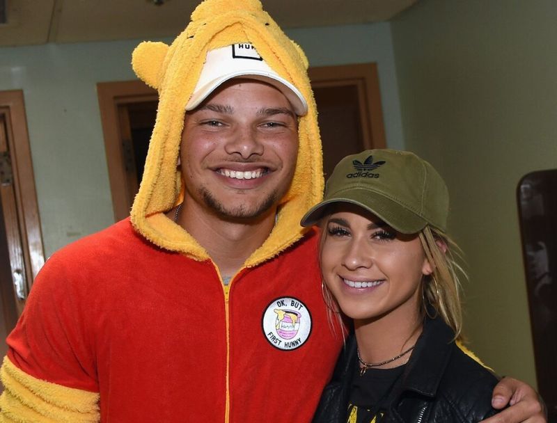 Kane Brown sonríe con un disfraz de Winnie the Pooh con su brazo alrededor de Katelyn Jae, sonriendo con una chaqueta de cuero y un sombrero verde