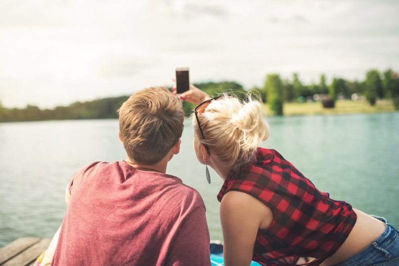 Imagen de una pareja tomándose una selfie juntos mientras están sentados en un muelle y mirando un lago.